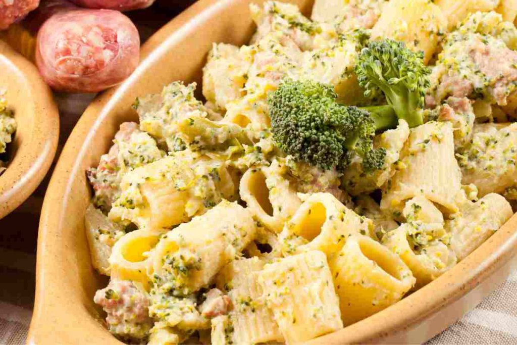 Pasta con broccoli e salsiccia in un piatto gratinato, con dietro una salsiccia intera