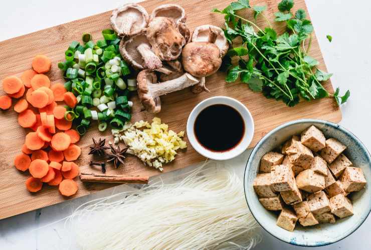 verdure e funghi sopra un tagliere con accanto una ciotola di tofu e una ciotolina di salsa di soia