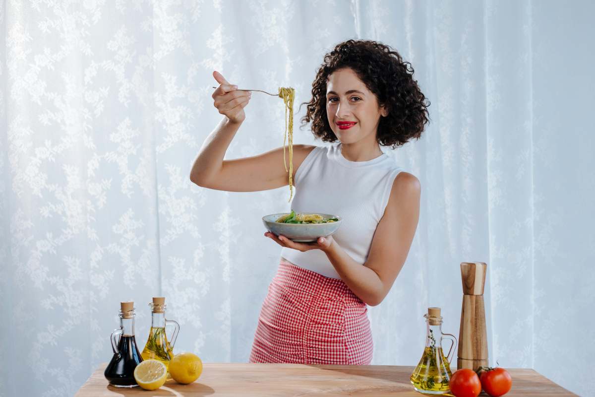 donna in piedi che mangia un piatto di pasta vicino bottigliette di olio e di aceto e un limone tagliato